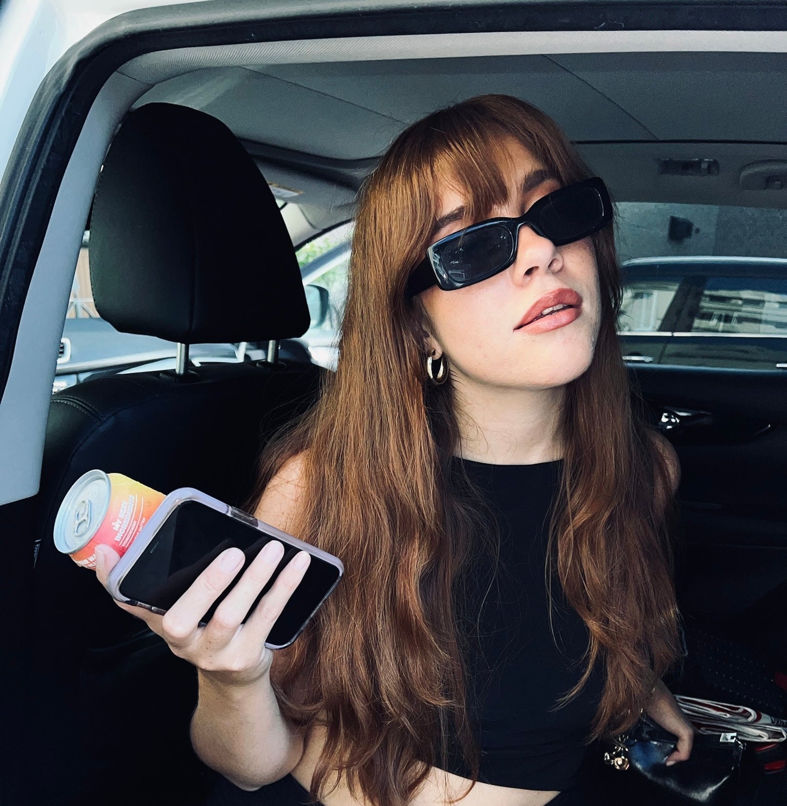 A woman with long brown hair, wearing black sunglasses and an all-black outfit, sits in a car holding a can of drink and a smartphone. The image evokes a stylish, casual vibe 