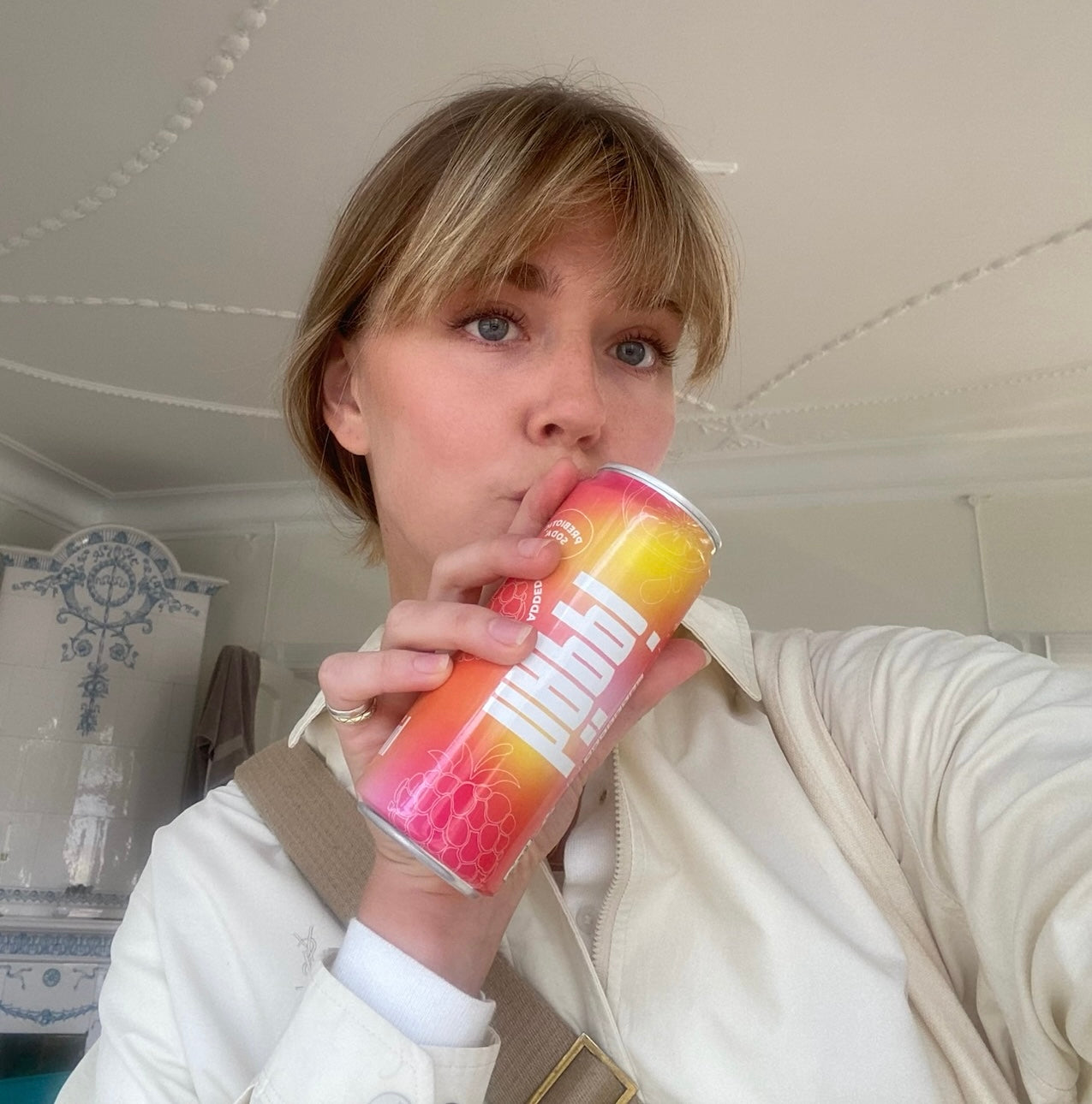 A woman enjoys a häppi prebiotic soda, capturing a refreshing and stylish moment indoors with a bright, fruity drink in hand.