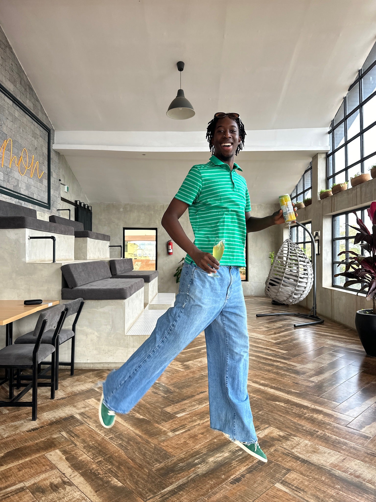 A cheerful person in a green-striped shirt and blue jeans holds a drink can, smiling mid-stride in a modern, well-lit café with large windows and cozy seating.