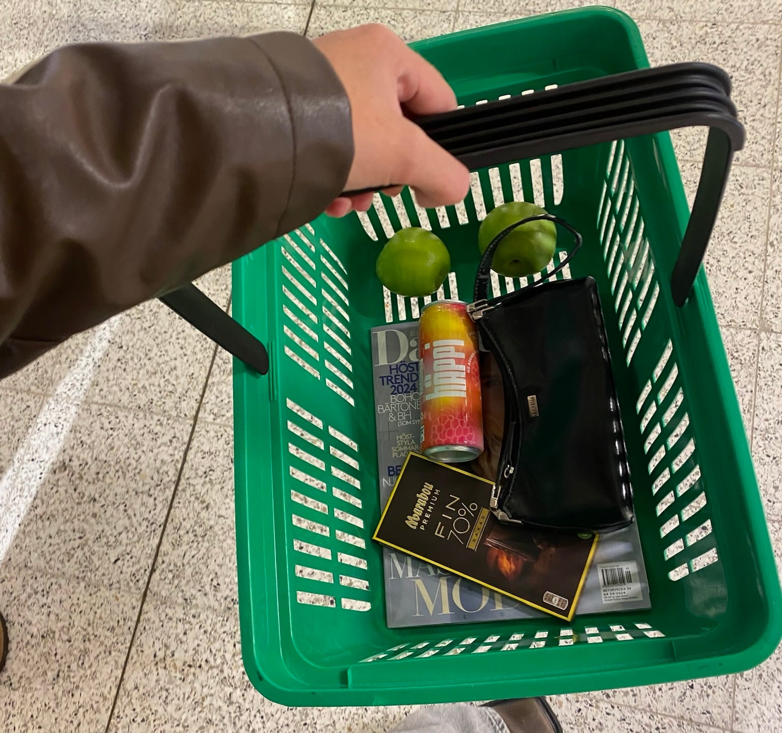 A green shopping basket featuring a hÄppi prebiotic soda, fresh green apples, a magazine, dark chocolate, and a black purse. 