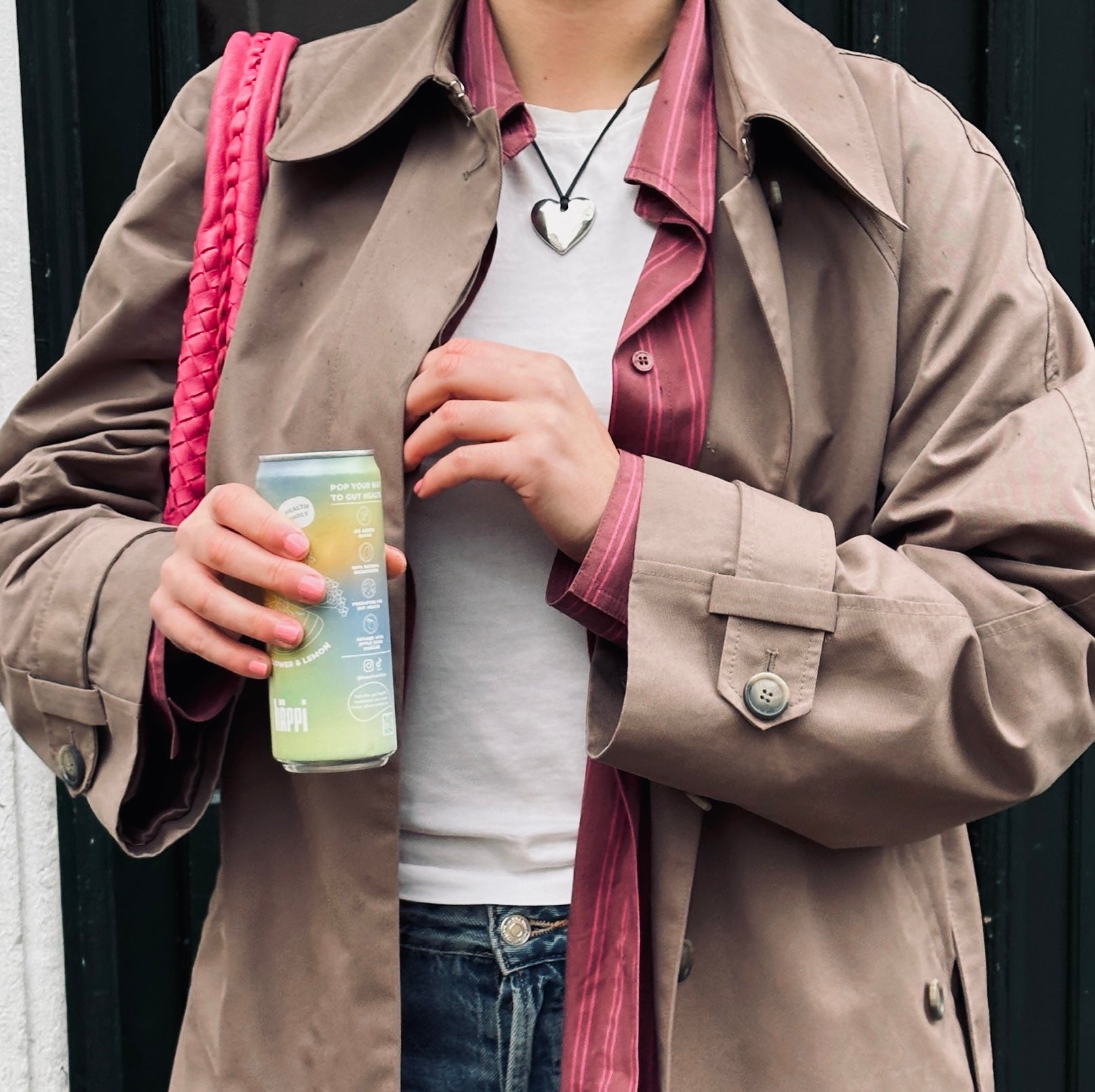 A person wearing a beige trench coat and a pink-striped shirt holds a can of hÄppi soda 