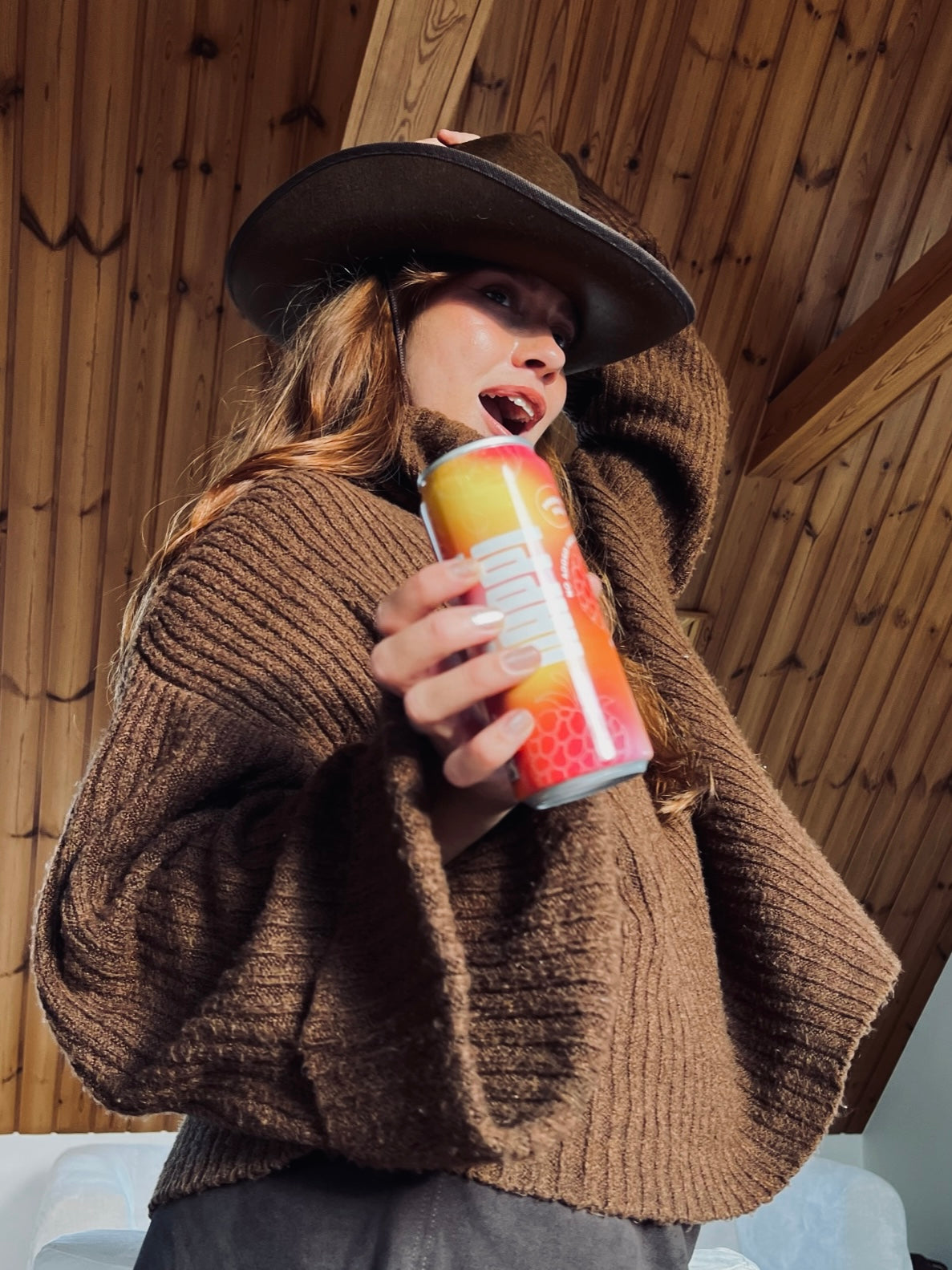 A woman in a cozy brown sweater and hat happily holds a vibrant hÄppi prebiotic soda can. The warm, rustic setting highlights the natural and wholesome vibe of the drink, perfect for health-conscious enjoyment.