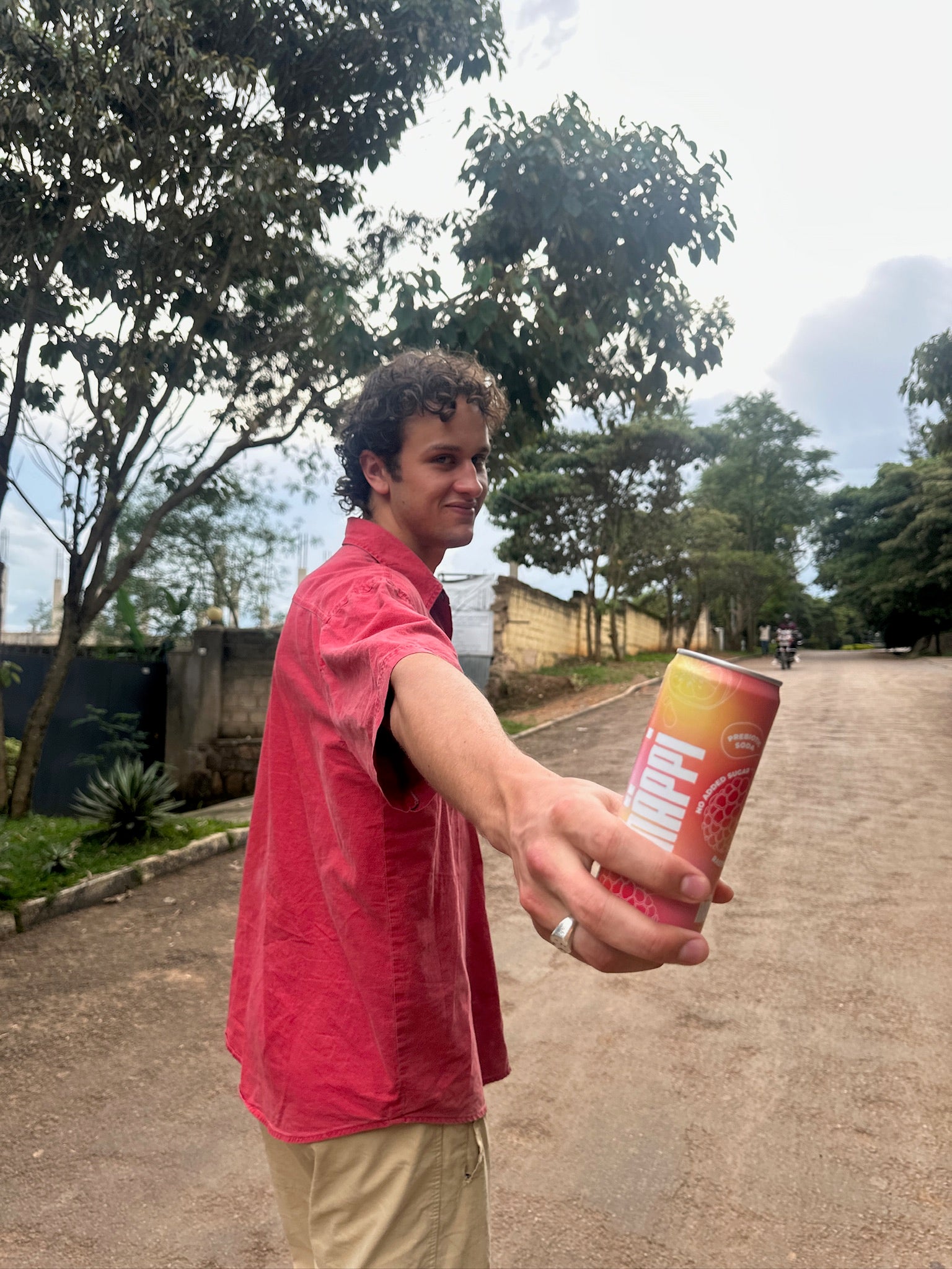 A young man in a red shirt offers a can of hÄppi soda while standing on a tree-lined street, smiling casually. The image conveys a relaxed, outdoor vibe.